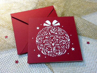 Rote Karte mit weiß ausgeschnittener Christbaumkugel, roter Umschlag auf goldfarbenem Netzstoff.