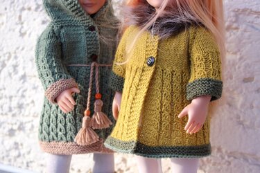Zwei Puppen in gestrickten Mänteln: grüner Mantel mit Quastenband und senfgelber Mantel mit Knopf und flauschigem Kragen