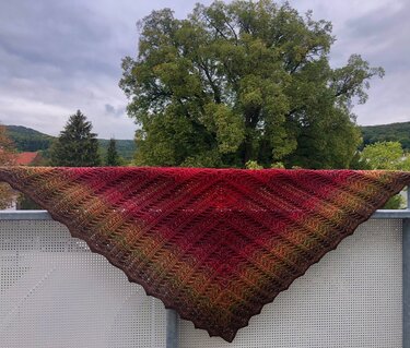 Gehäkeltes Dreieckstuch in Rot- und Grüntönen mit diagonalem Strukturmuster auf einem Balkongeländer vor Bäumen.