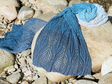 Blue ombre knitted lace shawl with openwork stitch pattern laid on grey rocks.