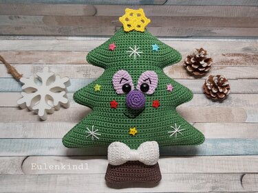 Gehäkelter grüner Amigurumi-Weihnachtsbaum mit Gesicht, gelber Sternspitze, weißen Sticksternen und weißer Schleife auf hellem Holzuntergrund, mit Tannenzapfen daneben.