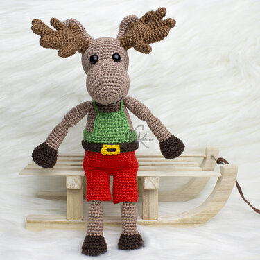 Handgearbeitetes, gehäkeltes Elch-Amigurumi mit braunem Geweih, grünem Oberteil, roter Hose und dunklen Füßen auf hölzernem Dekoschlitten