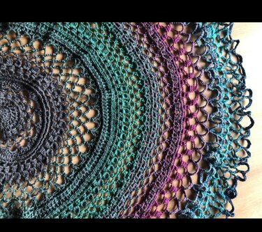 Nahaufnahme eines halbrunden Häkel­tuchs mit Farbverlauf von Grau über Türkis zu Violett und Hellblau sowie feinem Spitzenmuster