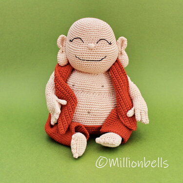Small beige crocheted figurine with a smiling face and reddish-orange vest seated on a green surface.