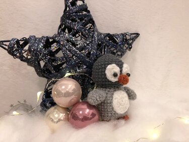 Gehäkelter Amigurumi-Pinguin in Grau und Weiß neben Weihnachtskugeln und sternförmiger Deko