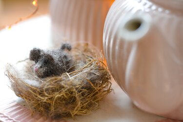 Graues Amigurumi-Mäuschen in einem Fasernest, neben hellen Keramikgefäßen.