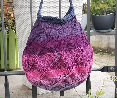 Gestrickte Ombre-Tasche in Blau, Violett und Pink mit Zopf- und Lochmustern, hängend an einem Metallgeländer.