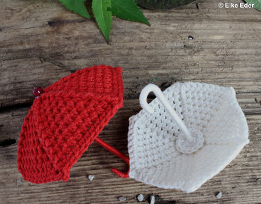 Overhead view of two small crocheted umbrella ornaments, one red and one white, on a wooden surface.