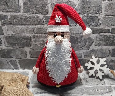 Gehäkelter Amigurumi-Weihnachtsmann mit roter Mütze und geschlungenem weißen Bart vor grauer Ziegelwand