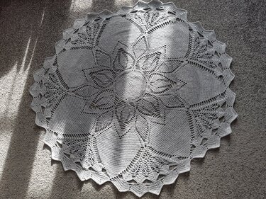 Overhead view of a large round grey crochet doily with intricate lace floral patterns on a carpet with sunlight.