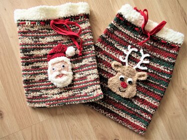 Zwei gehäkelte Nikolaussäckchen mit Weihnachtsmann- und Rentier-Applikationen, gestreiftes Rot‑Grün‑Creme-Muster, Zugband und flauschiger Rand auf Holzboden