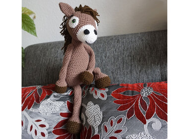 Brown crocheted amigurumi horse with dark mane and hooves, white muzzle, green eyes, on a red and grey floral cushion.