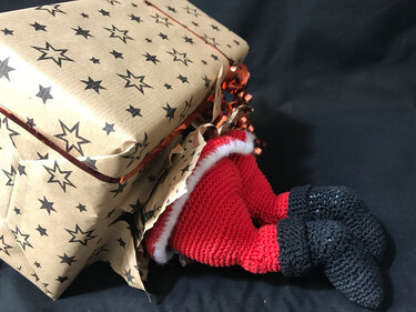 Gehäkelte Weihnachtsmann-Beine in Rot und Schwarz ragen aus Geschenk mit Sternenpapier
