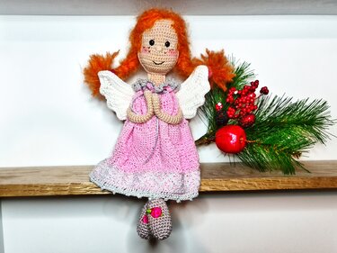 Gehäkelter Amigurumi-Weihnachtsengel in rosa Kleid mit weißen Flügeln und roten Zöpfen auf Regal neben Tannenzweig
