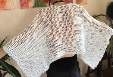Rückansicht einer Person in einem weißen, locker gehäkelten Poncho mit transparentem Lochmuster.