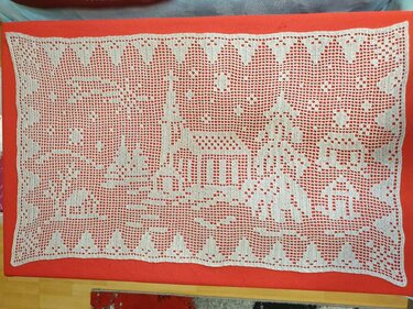 Weißer rechterckiger Tischläufer im Filet-Häkelmuster mit Dorfmotiv auf rotem Stoff.
