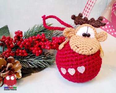 Crocheted reindeer ornament with a red round body, beige face, brown antlers and three iridescent heart appliqués, shown with red berries and an evergreen sprig.