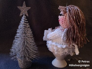 Weiß gehäkelter Engel-Eierwärmer mit braunen Garnhaaren auf einem weißen Eierbecher neben silbernem Deko-Tannenbaum