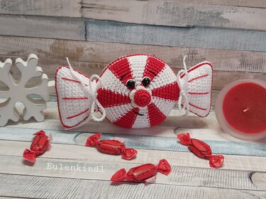 Gehäkeltes rot-weiß gestreiftes Bonbon mit Gesicht, umgeben von kleineren Süßigkeiten, roter Kerze und Schneeflocken-Deko auf hellem Holztisch.