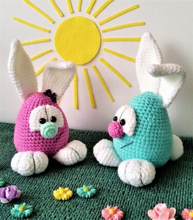 Zwei gehäkelte Amigurumi-Hasen in Rosa und Mint mit weißen Ohren und Pfoten vor einer gelben Sonnendekoration