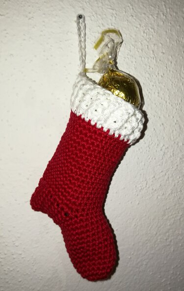 Roter gehäkelter Weihnachtsstrumpf mit weißem Bündchen an einer Wand hängend, goldverpackte Süßigkeit ragt heraus
