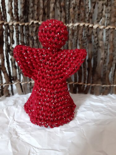 Gehäkelter roter Engel mit silbernem Glitzer vor rustikaler Holzwand auf weißem, zerknittertem Untergrund.