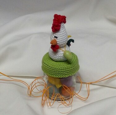 Glas mit grünem Häkeldeckel, darauf eine weiße gehäkelte Henne mit rotem Kamm; Glas gefüllt mit pastellfarbenen Eiern und oranger Bastdeko