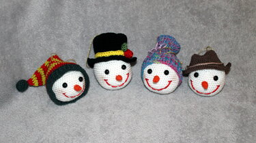 Vier gehäkelte Schneemann-Anhänger mit Schlaufen und unterschiedlichen Hüten auf grauem Stoff