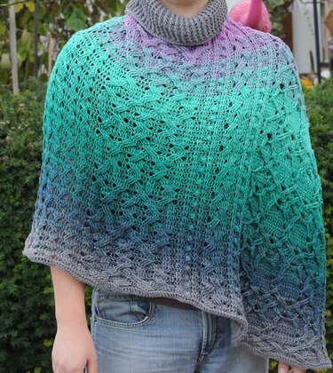 Person trägt gehäkelten Poncho mit Farbverlauf von Grau zu Türkis und Violett sowie grauem Rollkragen.