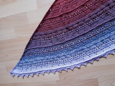 Gehäkeltes dreieckiges Tuch mit Farbverlauf von Rot über Lila zu Hellblau auf hellem Holzboden