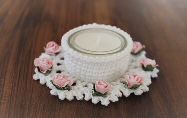 Weißer gehäkelter Teelichthalter mit rosa Rosen und Teelicht im Glas auf dunklem Holz.