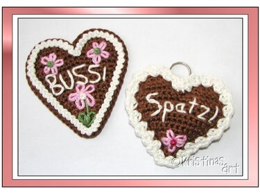 Zwei gehäkelte herzförmige Schlüsselanhänger in Braun mit weißer Bordüre, rosa Blumen und aufgestickten Schriftzügen