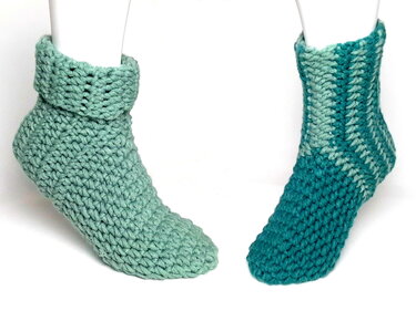 Zwei gehäkelte Socken in Grün- und Türkistönen auf weißen Mannequin-Füßen, eine mit umgeschlagenem Bündchen.