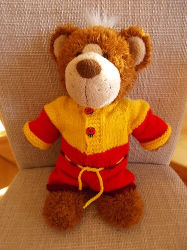 Teddyb
R mit gelb-rotem, gestricktem Zweiteiler, Marienk
Rferkn
pfen und gelbem Kordelzug auf einem Stuhl