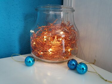 Transparentes Glasgefäß mit kupferfarbenem Lametta, Lichterkette und ausgeschnittener Tannenbaum-Silhouette, davor drei türkisblaue Weihnachtskugeln.