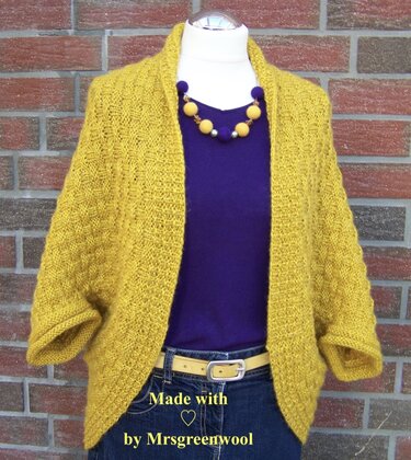 Mannequin trägt senfgelben, texturierten Strick-Seelenwärmer mit breitem Kragen über dunklem Top und Jeans vor Backsteinwand