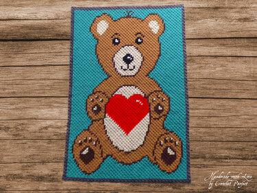 Gehäkelte Babydecke im Pixelmuster mit sitzendem braunem Teddybär, rotem Herz auf türkisfarbenem Grund und lila Rand, auf Holzuntergrund