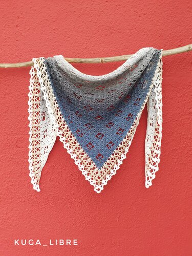 Crocheted triangle shawl in blue-gray gradient with cream and white lace border hanging on a wooden branch against a red wall