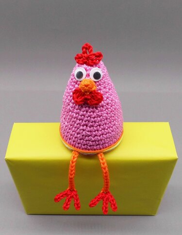 Rosa gehäkeltes Huhn mit Wackelaugen und orangen Beinen auf gelbem Kasten