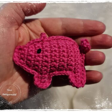 Hand mit kleinem, flachen rosa gehäkelten Glücksschwein mit schwarzen Augen und Ringelschwanz