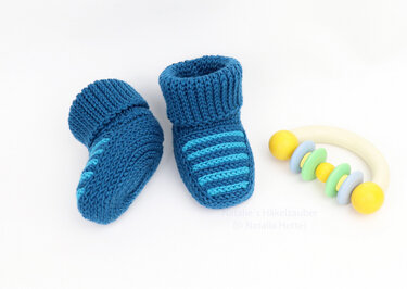 pair of blue crocheted baby booties with ribbed cuffs and light-blue stripes beside a pastel rattle