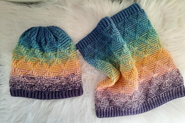 Mütze und Loop‑Schal mit Farbverlauf von Blau über Gelb zu Violett, strukturiertes Korbmuster, auf hellem Felluntergrund.