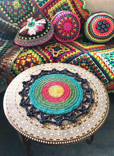 Boho-Häkelarrangement: rundes, buntes Deckchen auf Tisch vor gehäkelter Decke und bunten Kissen