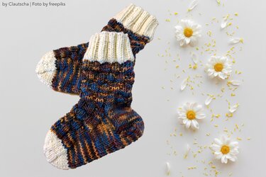 Gestrickte Kindersocken aus meliertem Garn mit cremefarbenen Zehen und Rippbündchen auf hellem Hintergrund neben Gänseblümchen