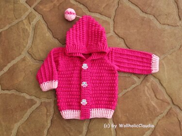 Fuchsiafarbene gehäkelte Babyjacke mit Kapuze und Eulenknöpfen auf braunem Steinboden
