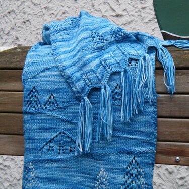 Variegated blue knitted scarf with lace triangle motifs and tassels on a wooden bench.