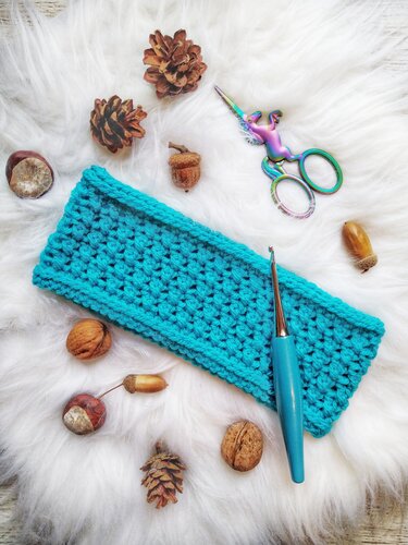 Teal crocheted headband on white faux fur with pinecones, acorns, crochet hook, and decorative scissors.