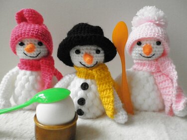Drei gehäkelte Schnemann-Eierwärmer mit schwarzer, pinker und rosafarbener Mütze, davor ein Ei im Eierbecher