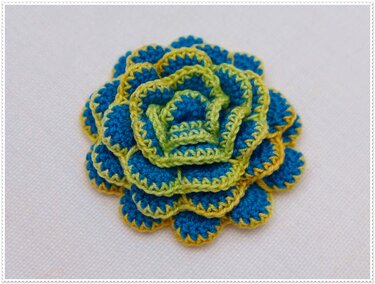 Mehrlagige gehäkelte Blütenapplikation in Blau mit lindgrünen und gelben Rändern auf hellem Stoff.