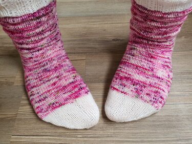 Zwei gestrickte Socken in Pink und Weiß auf Holzfußboden, Nahaufnahme der Zehen und Bündchen.